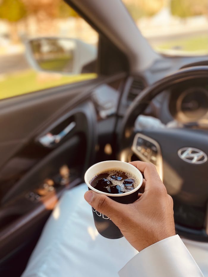 A person holding a hot beverage in a disposable cup inside a parked car, enjoying a peaceful morning.